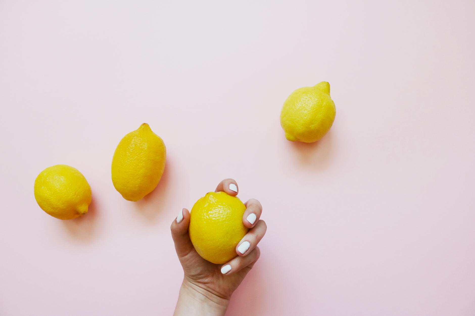 A hand holding a lemon on a soft pink background with additional lemons, representing fresh pleasure and natural arousal