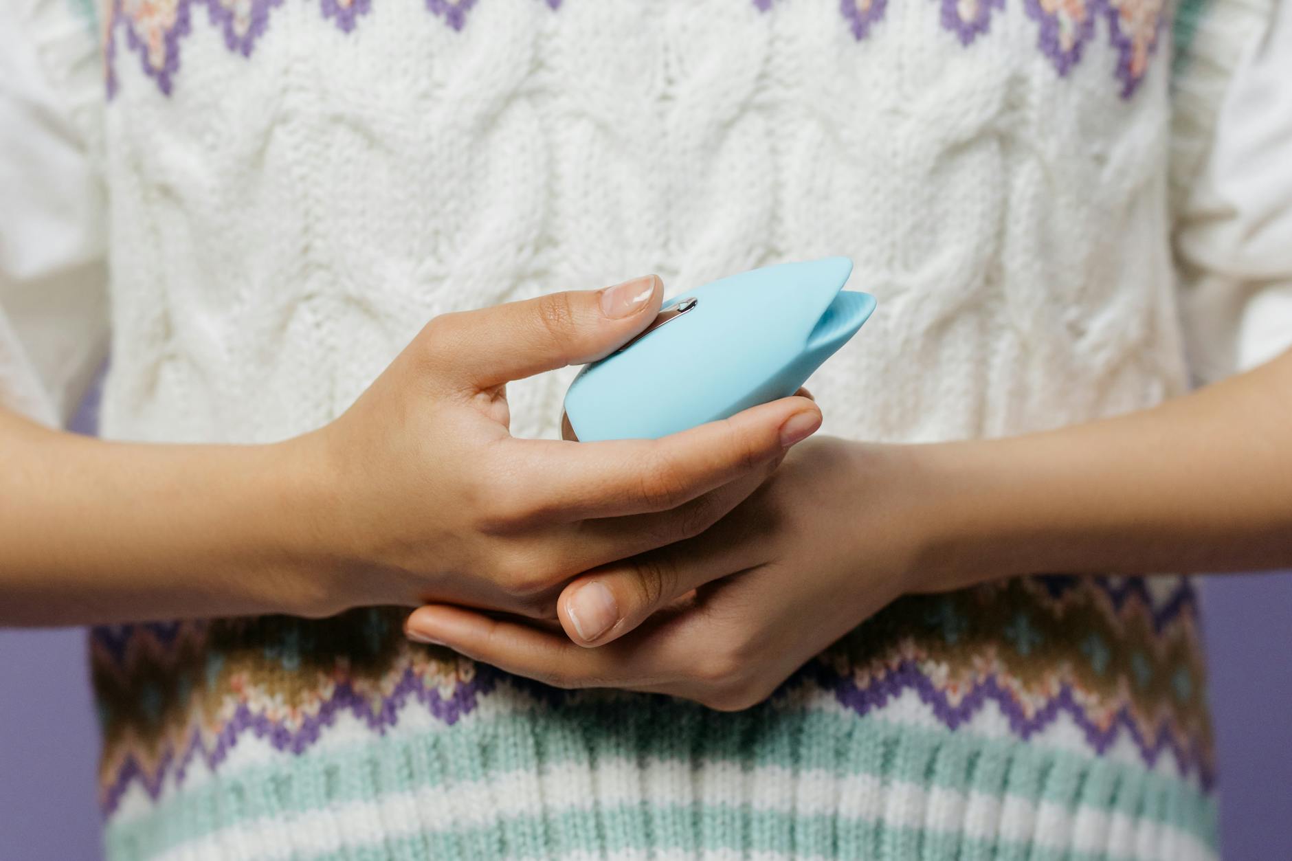 Detalhe das mãos segurando um vibrador de cor azul contra um suéter texturizado
